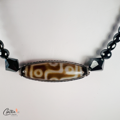 Necklace featuring an ancient two-eye Dzi bead from Nepal, paired with handcrafted pyramid-shaped ceramic beads and egg-shaped hematite beads, 15 inches with a 2-inch extender.
